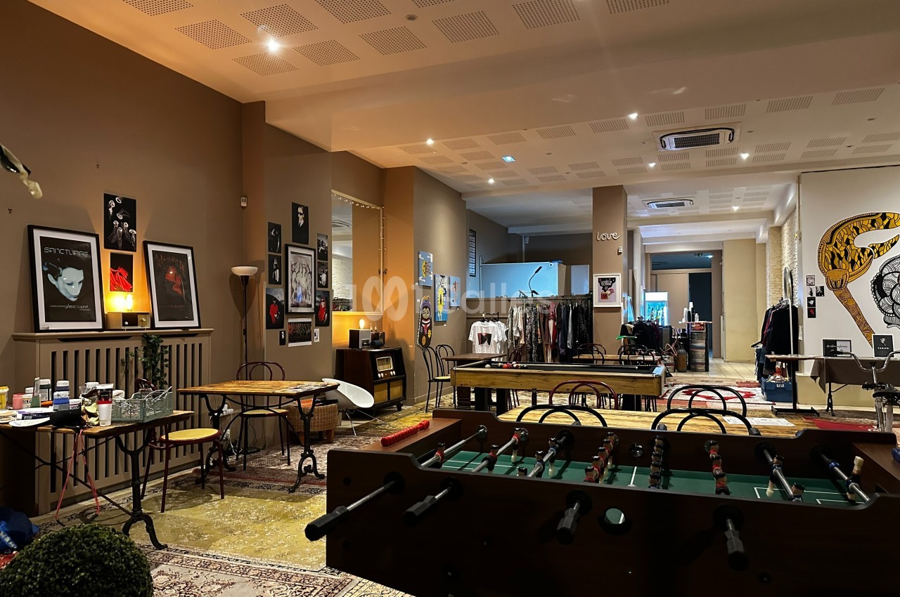 Salle aménagée avec des tables, un baby-foot, des affiches aux murs et des vêtements exposés en arrière-plan.