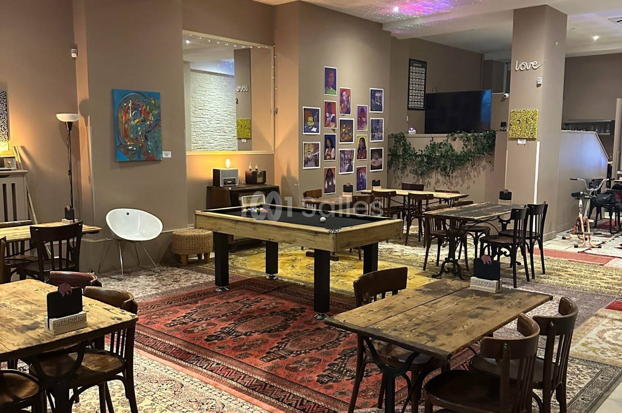Salle conviviale avec tables en bois, tapis colorés, un billard central et des décorations murales variées.