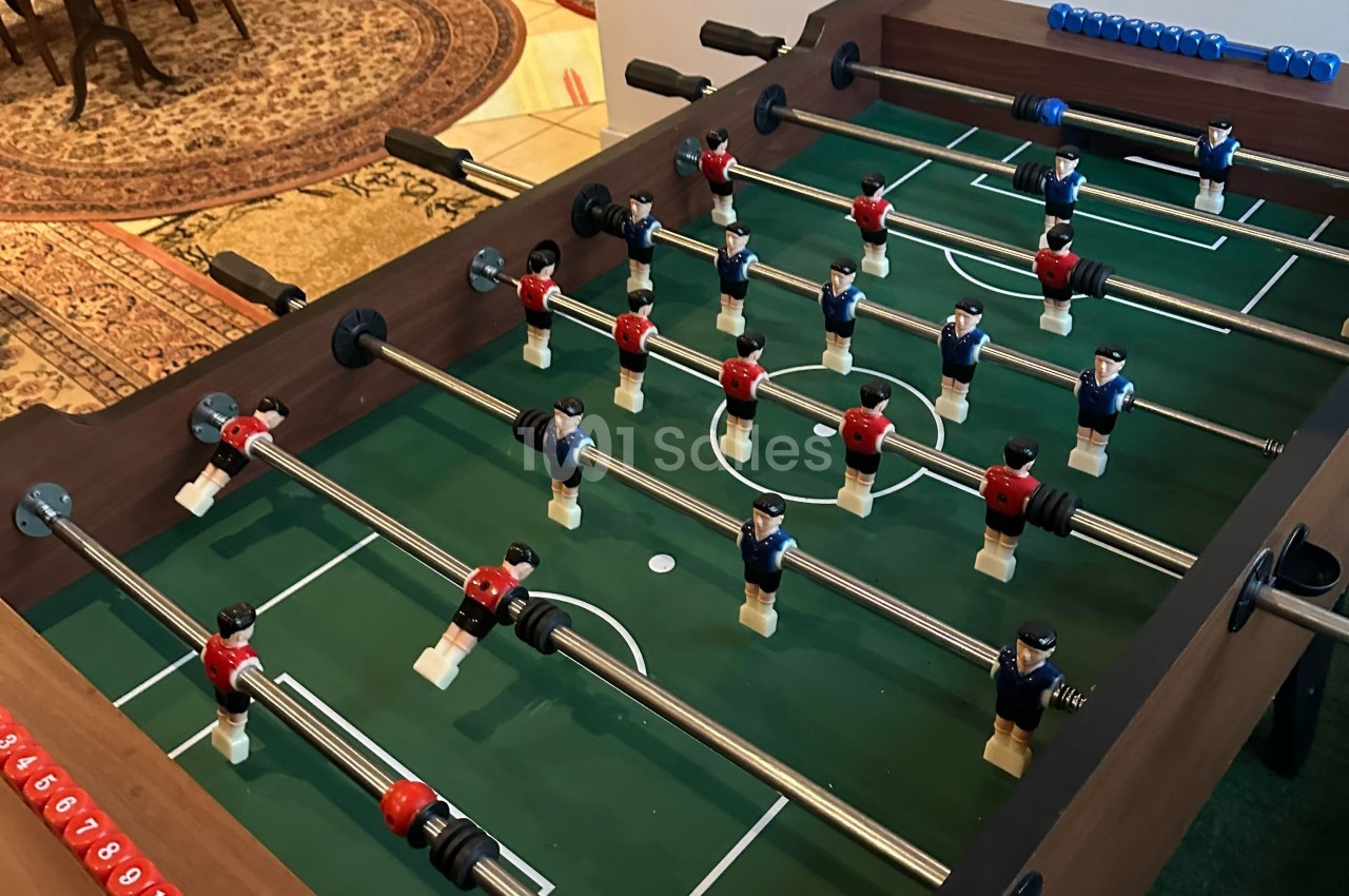 Table de baby-foot avec des joueurs rouges et bleus alignés sur un terrain vert, située dans un espace intérieur.