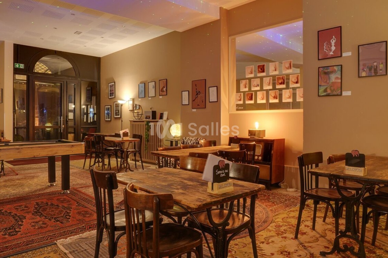 Salle chaleureuse avec tables en bois, tapis colorés, billard, et murs décorés de cadres artistiques.