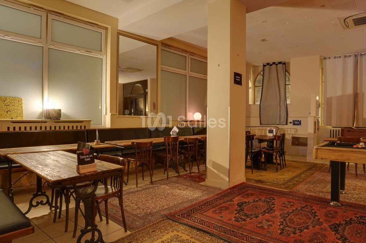 Salle avec tables en bois, chaises, banquettes, tapis colorés au sol et baby-foot dans un espace lumineux.