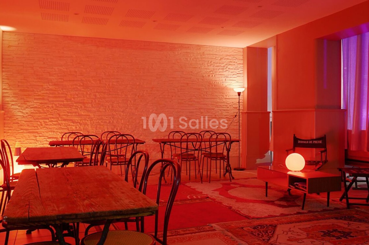 Salle avec tables en bois, chaises noires, murs en briques blanches et éclairage rouge tamisé.