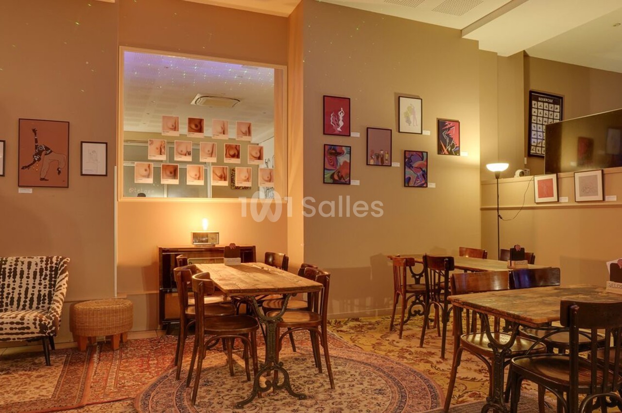 Salle de café chaleureuse avec tables en bois, chaises anciennes, murs décorés de cadres et éclairage tamisé.