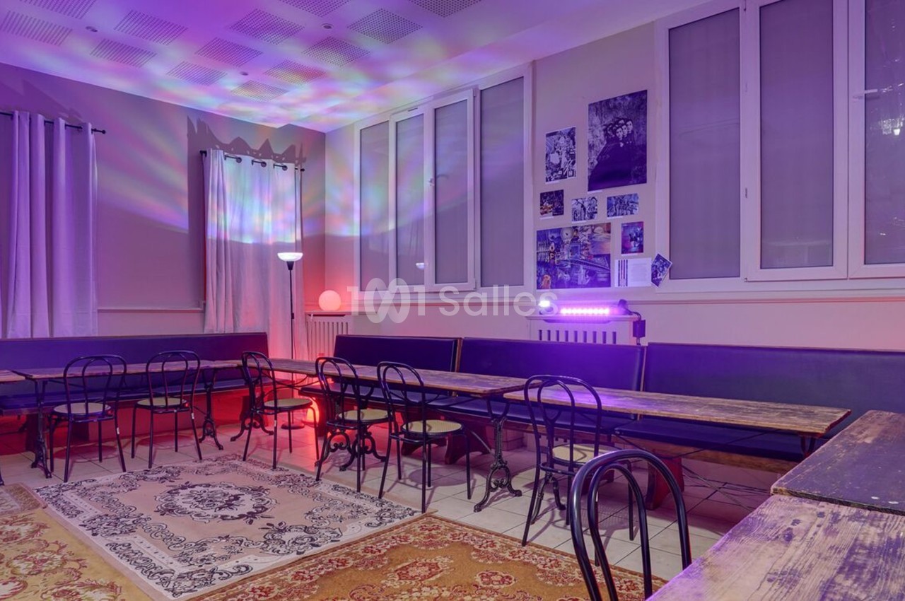 Salle avec des tables en bois, chaises en métal, tapis au sol et éclairage coloré diffus.