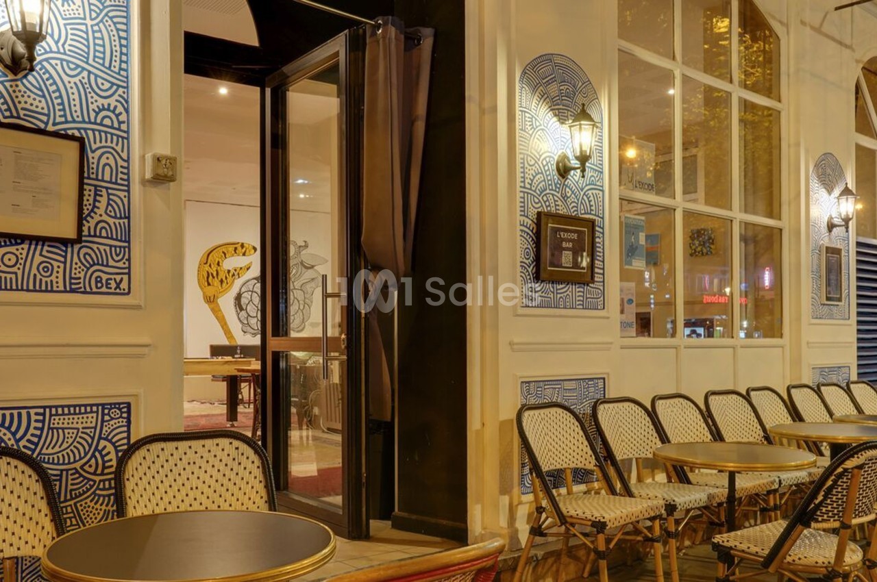 Façade d'un café avec terrasse extérieure, chaises en osier et tables rondes alignées, éclairage chaleureux en soirée.