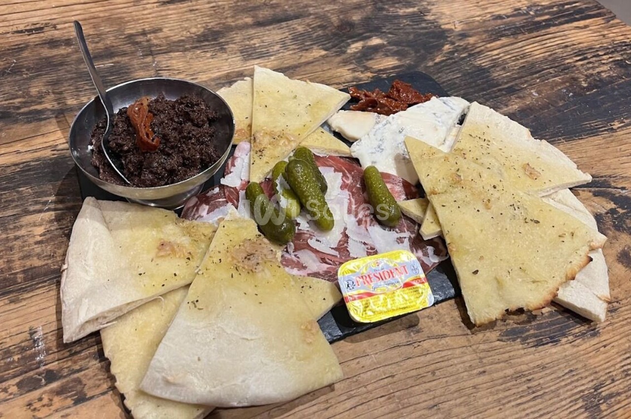 Assiette de charcuterie avec pain, cornichons, tapenade, fromage et beurre, présentée sur une table en bois.