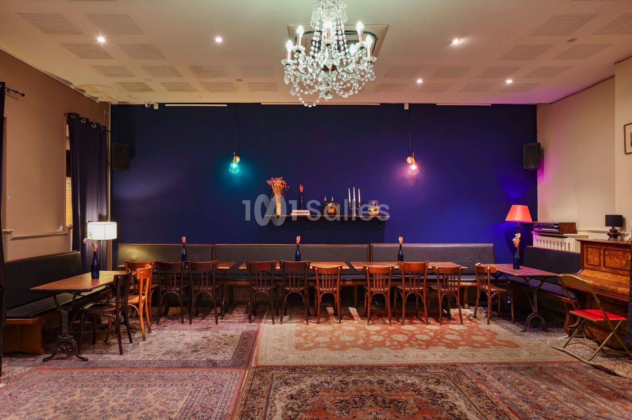 Salle avec tables et chaises alignées, murs bleu foncé, tapis au sol et éclairage chaleureux avec lustre au plafond.