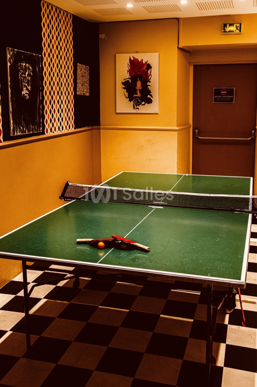 Table de ping-pong avec deux raquettes et une balle, située dans une pièce au sol à carreaux noir et blanc.