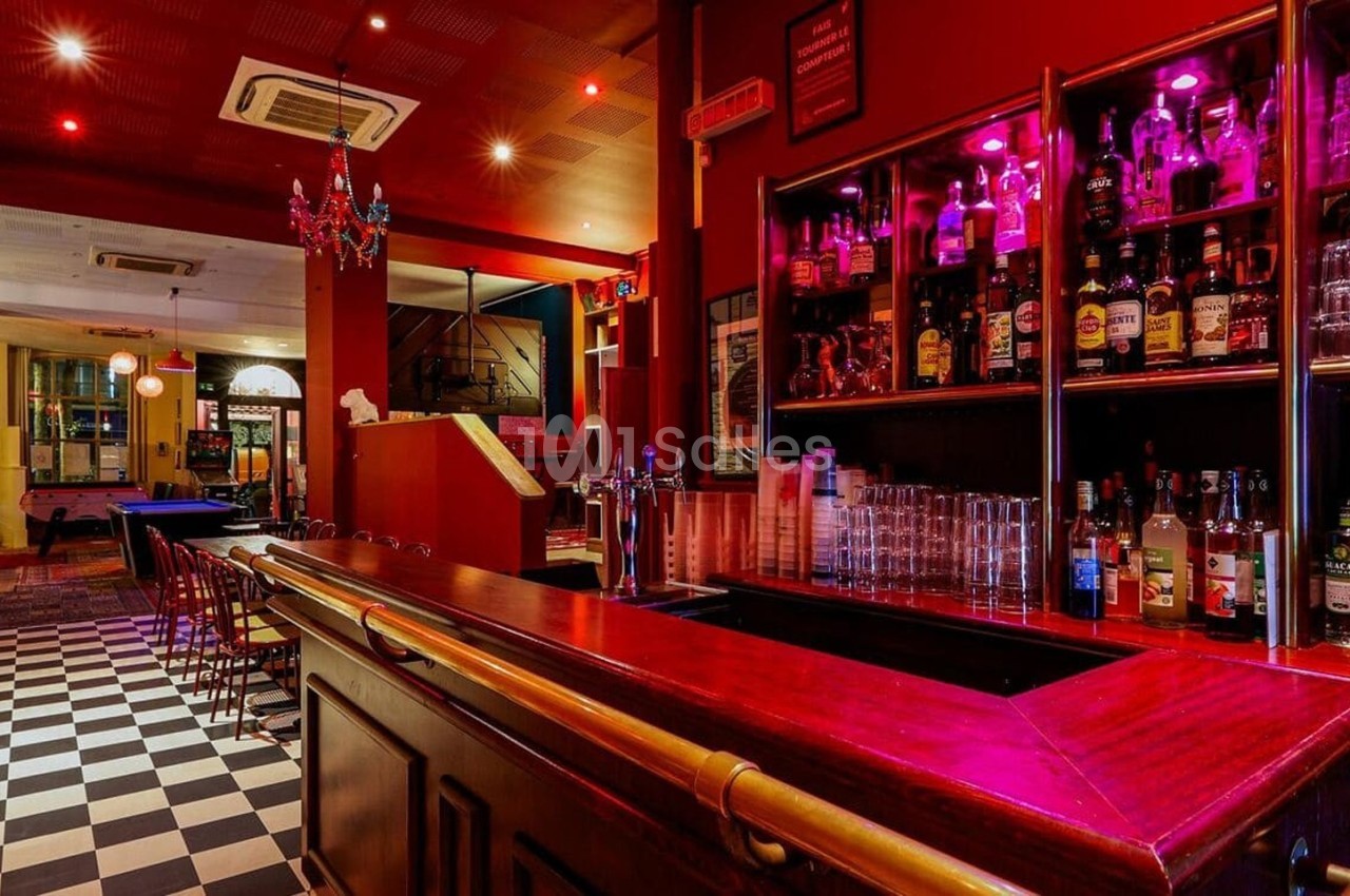 Bar avec comptoir en bois, étagères remplies de bouteilles, éclairage rouge et sol en damier noir et blanc.