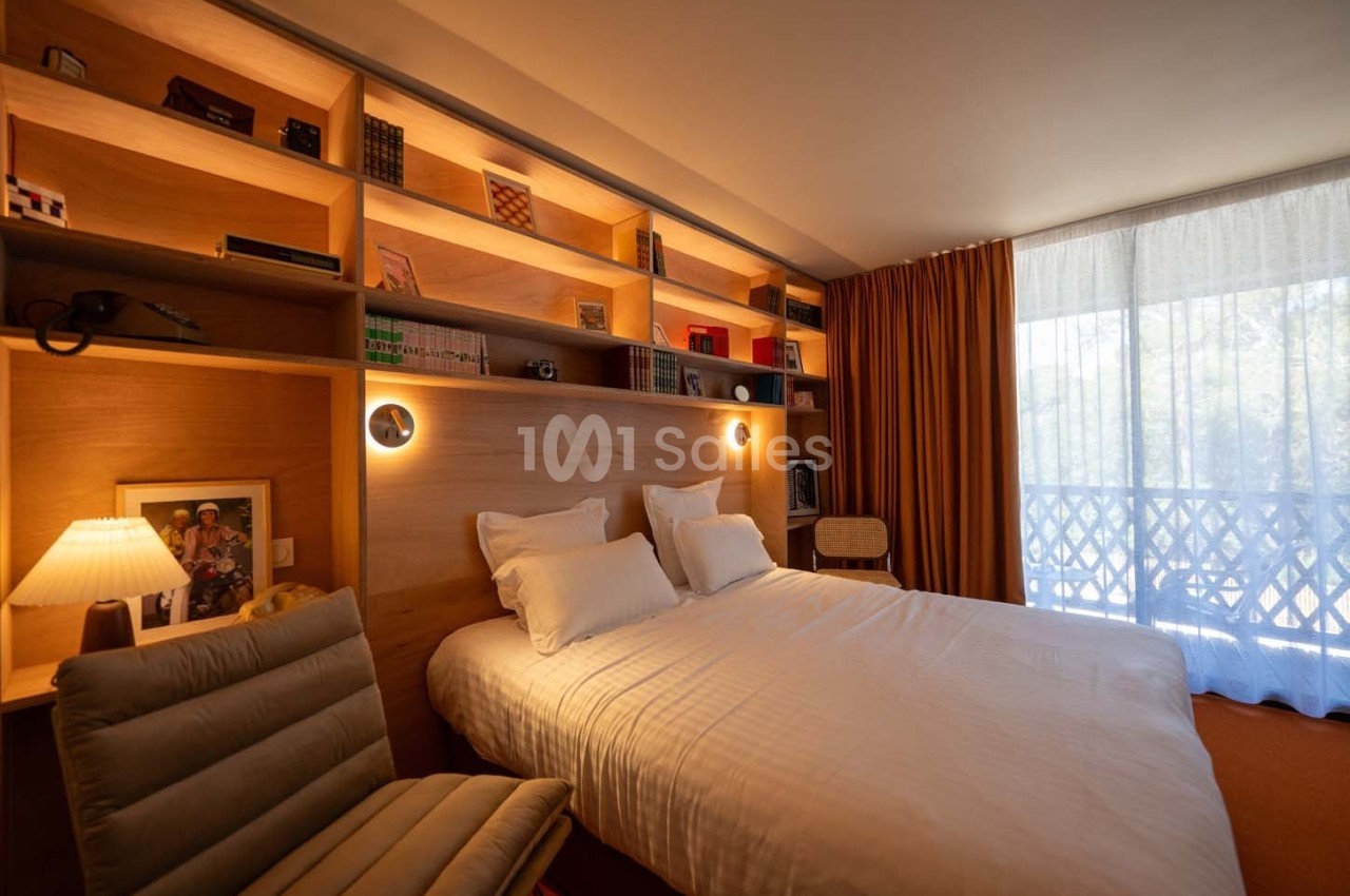 Chambre lumineuse avec lit double, étagères en bois remplies de livres et objets, et grande fenêtre avec rideaux.