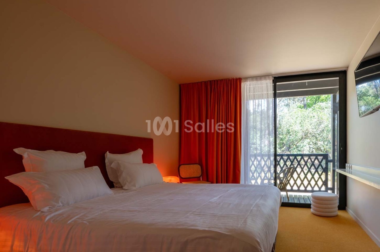 Chambre lumineuse avec lit double, rideaux rouges, balcon donnant sur un espace vert et mobilier minimaliste.