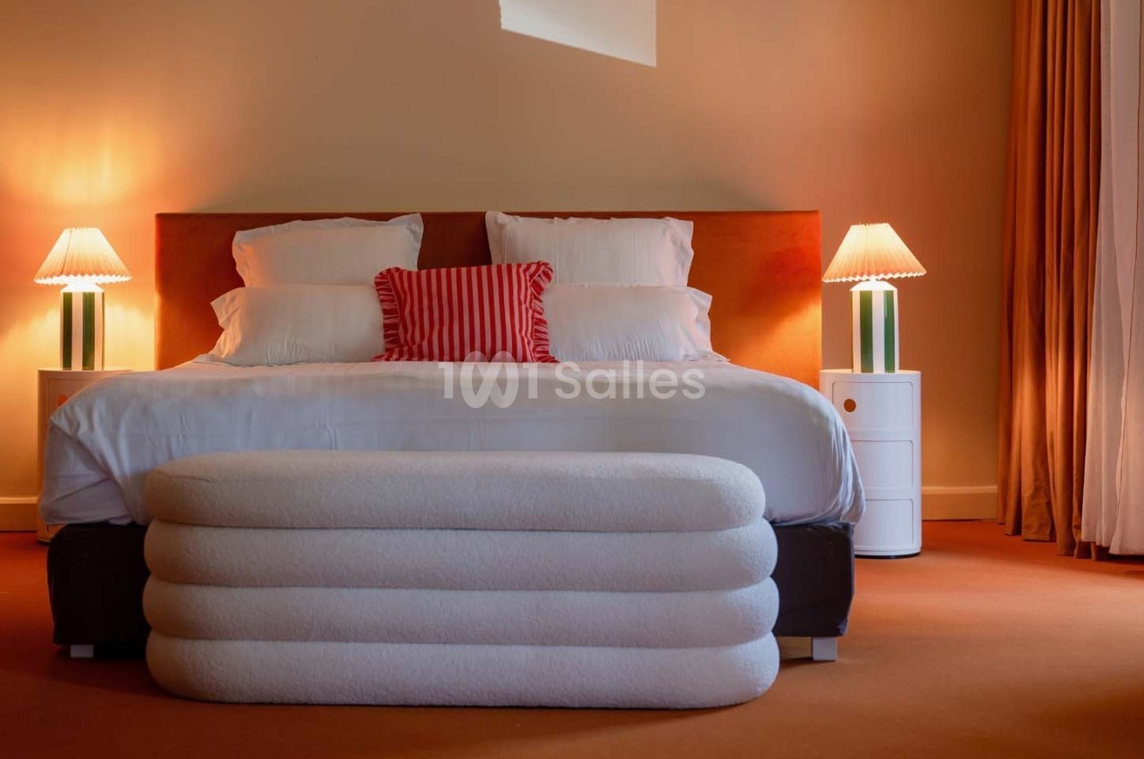 Lit double avec tête de lit orange, draps blancs, coussin rayé rouge, lampes de chevet et tabouret rembourré devant.