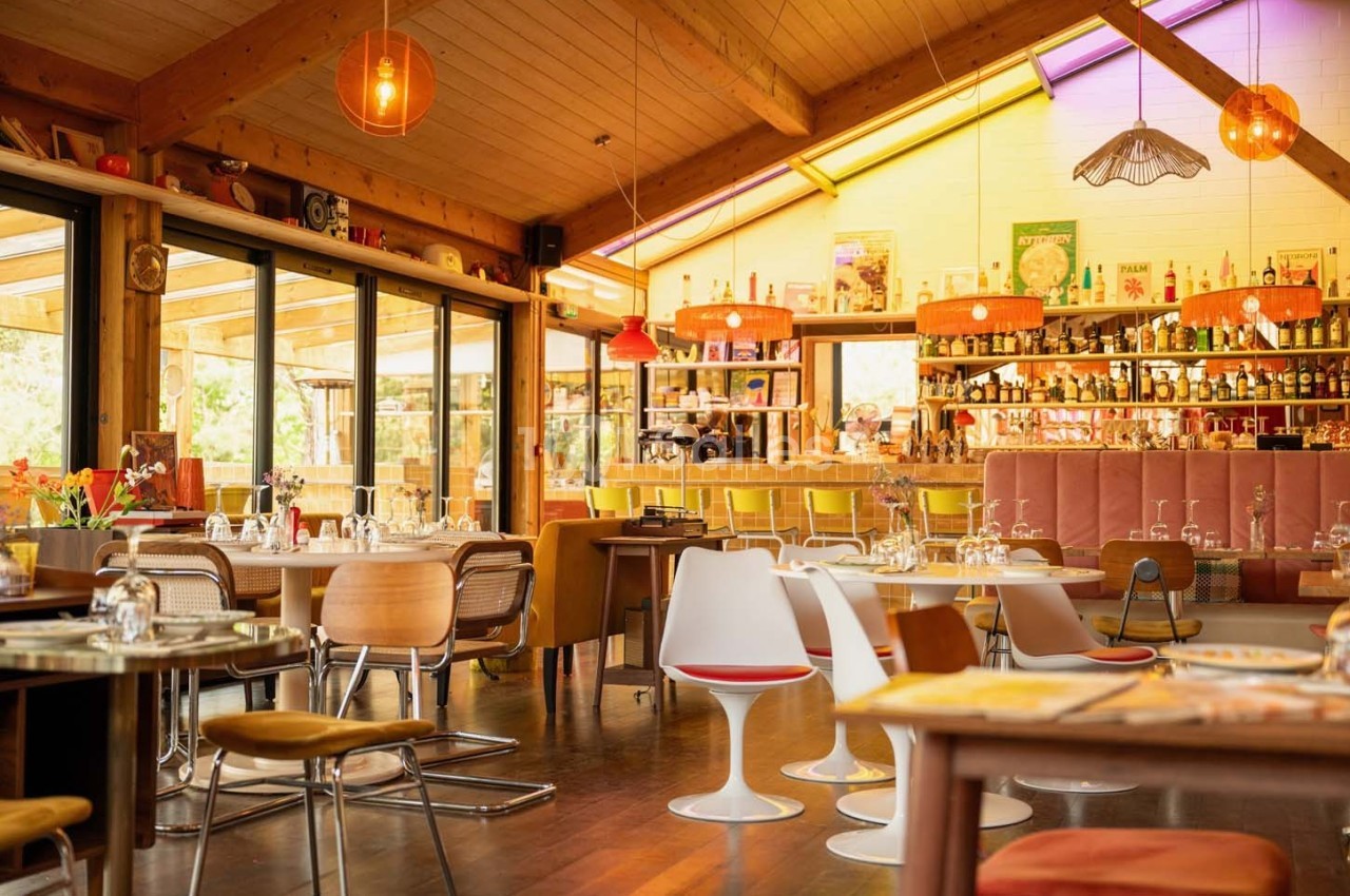 Salle de restaurant lumineuse avec mobilier coloré, grandes baies vitrées et bar en arrière-plan.