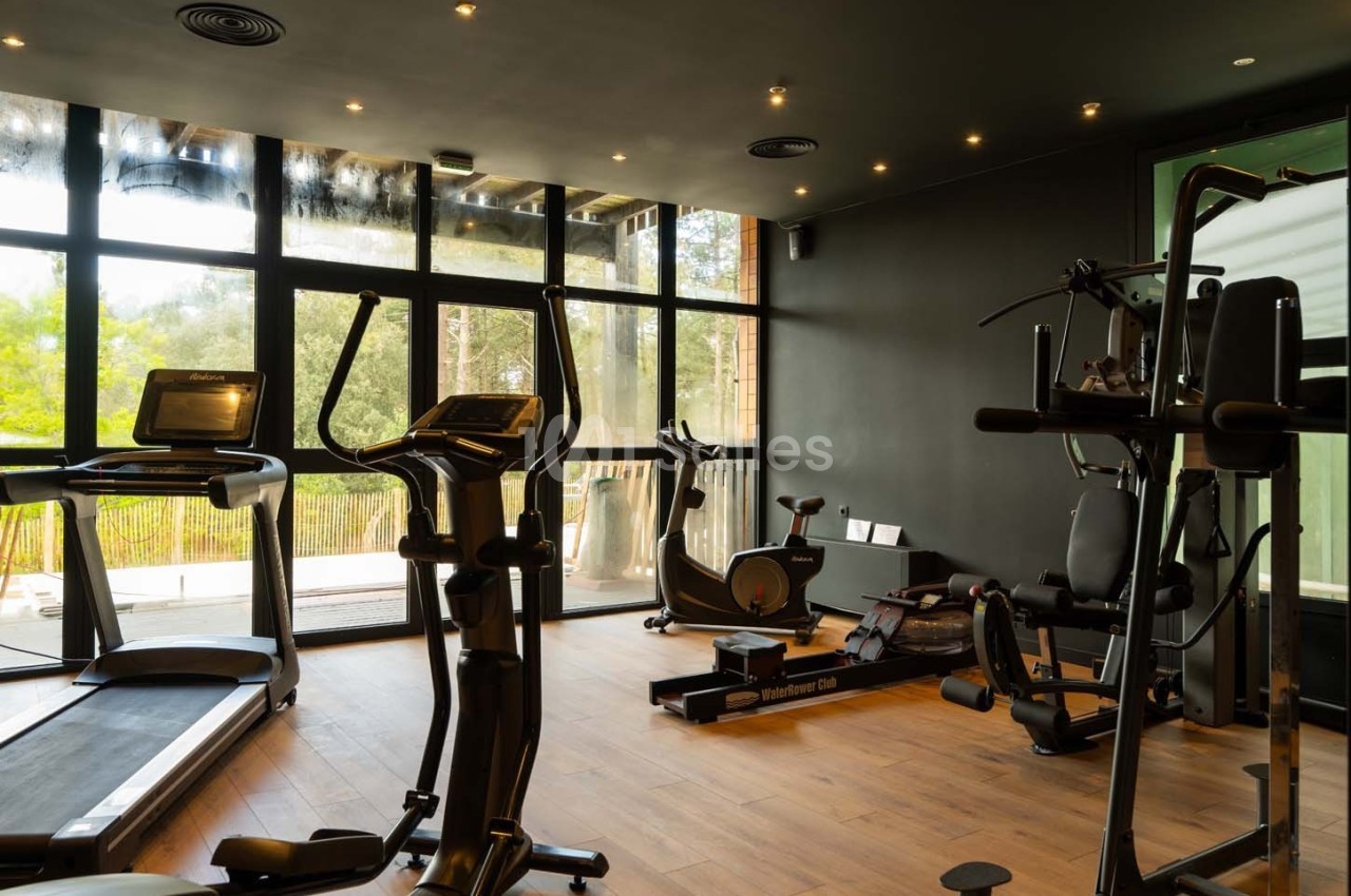 Salle de sport équipée de tapis de course, vélos d'exercice et machines de musculation, avec vue sur l'extérieur.
