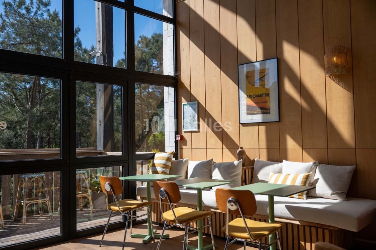 Salle lumineuse avec grandes baies vitrées, tables en bois, chaises jaunes et banquette avec coussins rayés.