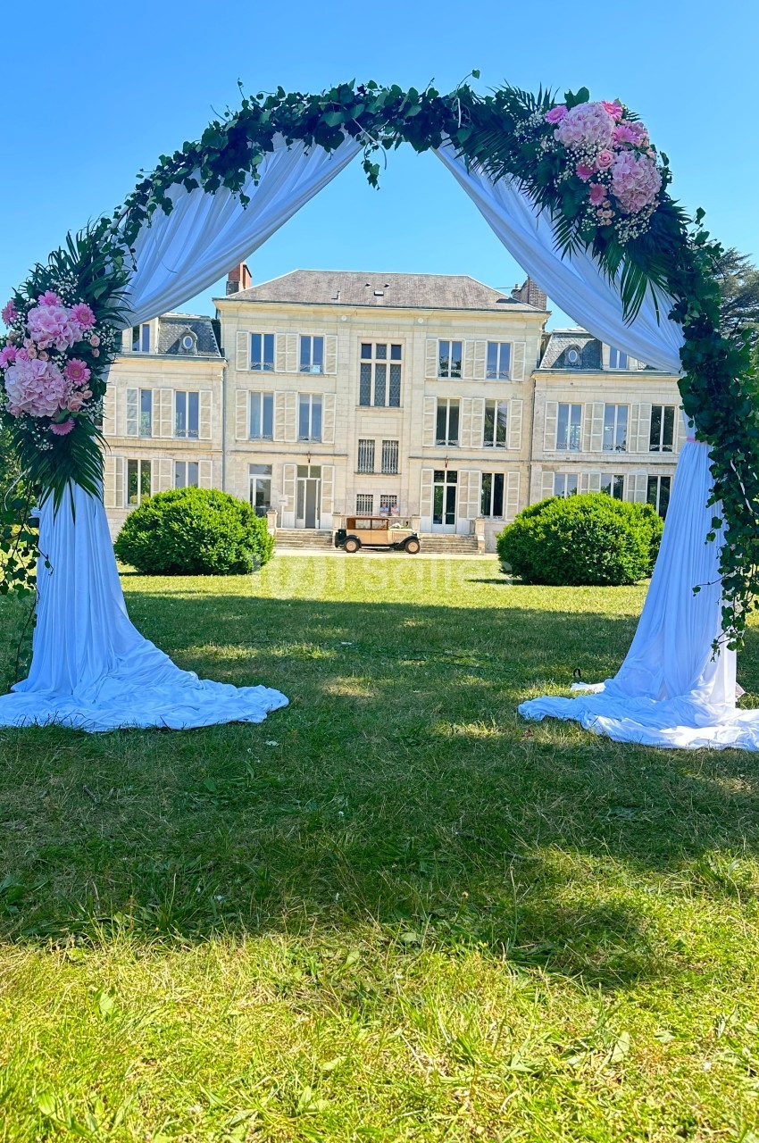 Arche de mariage décorée de fleurs et de drapés blancs, installée devant un château sous un ciel bleu.