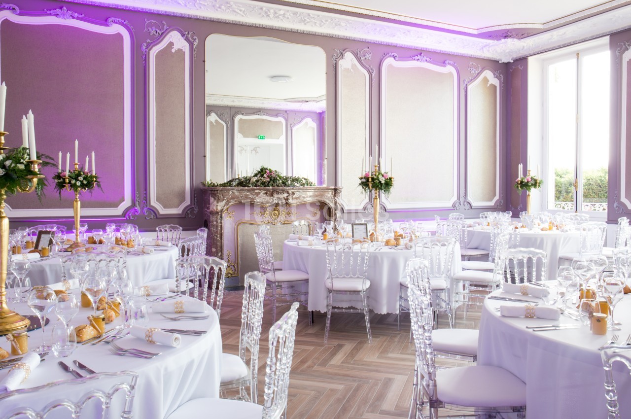 Salle de réception élégante avec tables dressées, chaises transparentes et décoration florale, éclairée par une lumière…