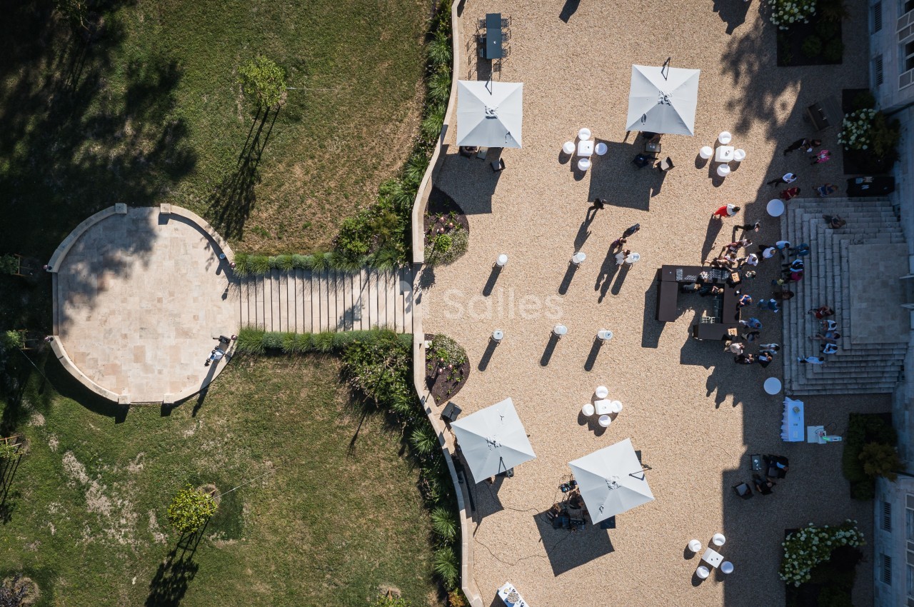 Vue aérienne d'une terrasse avec des parasols blancs, des tables et des personnes, entourée de pelouse et d'escaliers.