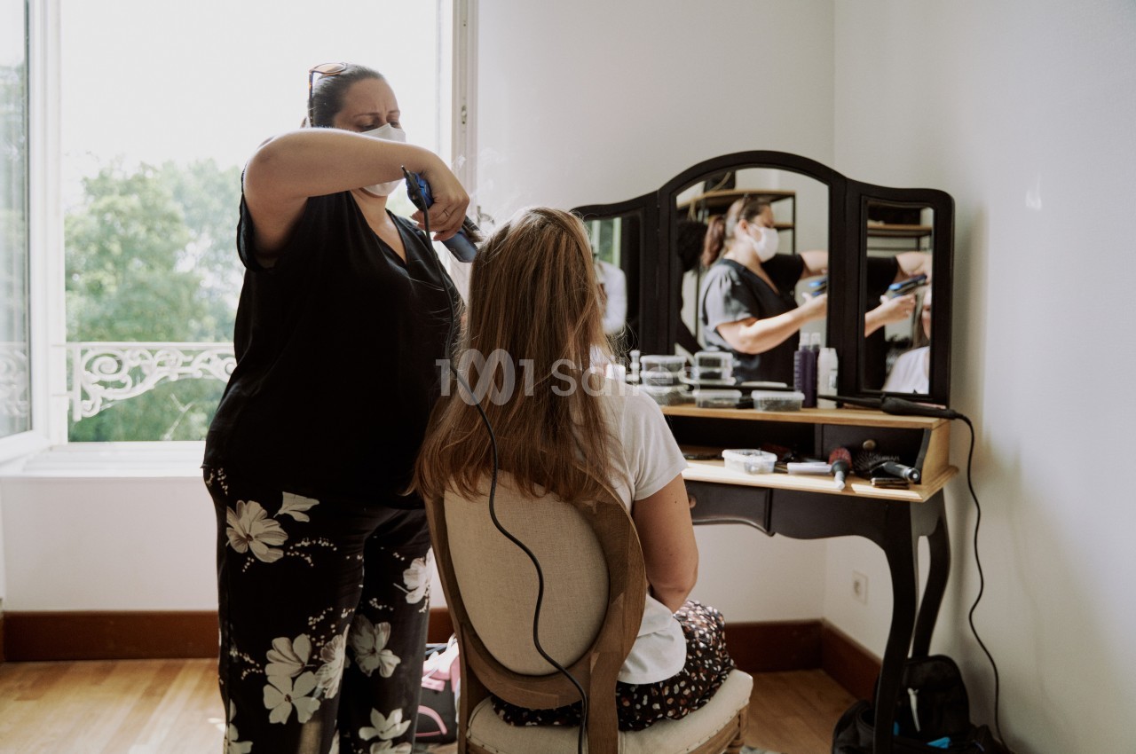 Une coiffeuse stylise les cheveux d'une femme assise devant une coiffeuse dans une pièce lumineuse.