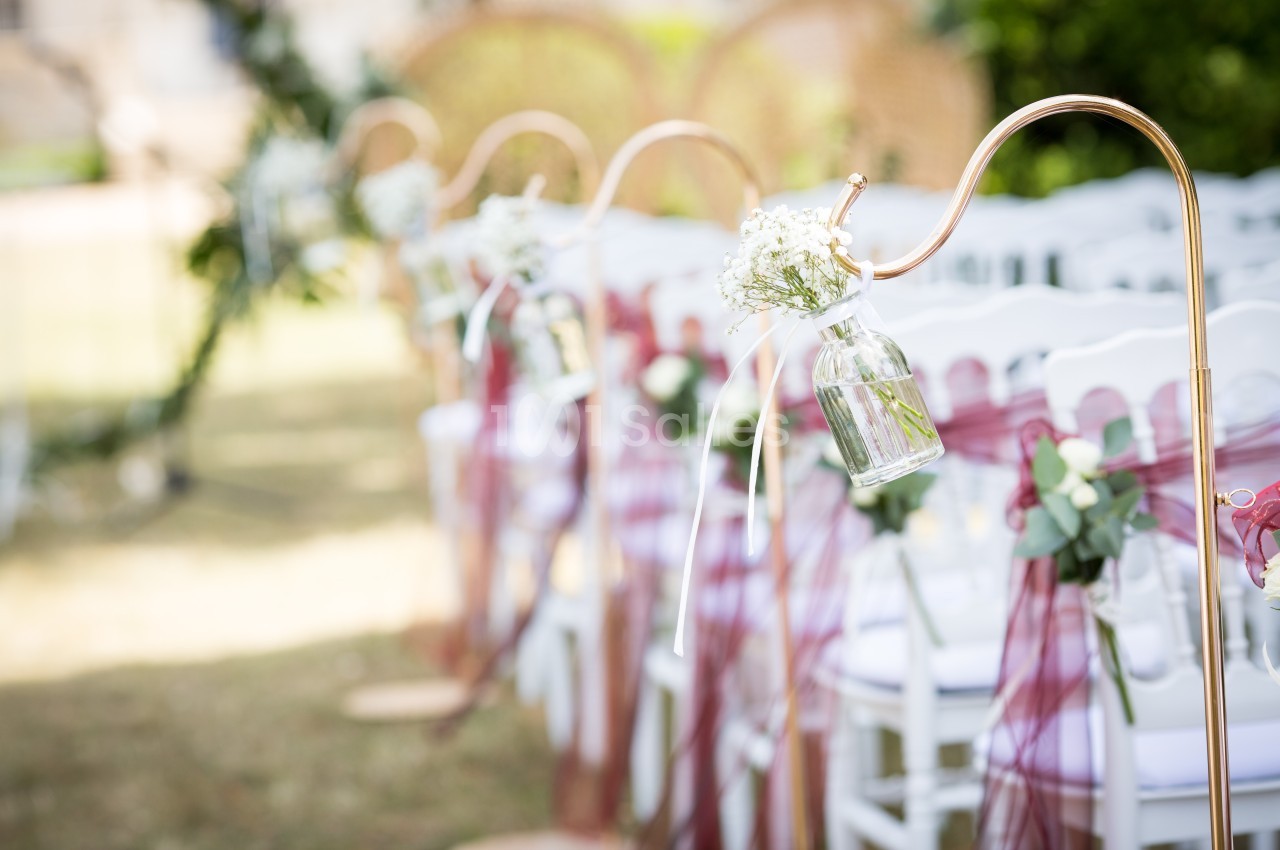 Chaises blanches alignées avec décorations florales suspendues dans des vases en verre, lors d'une cérémonie en extérieur.