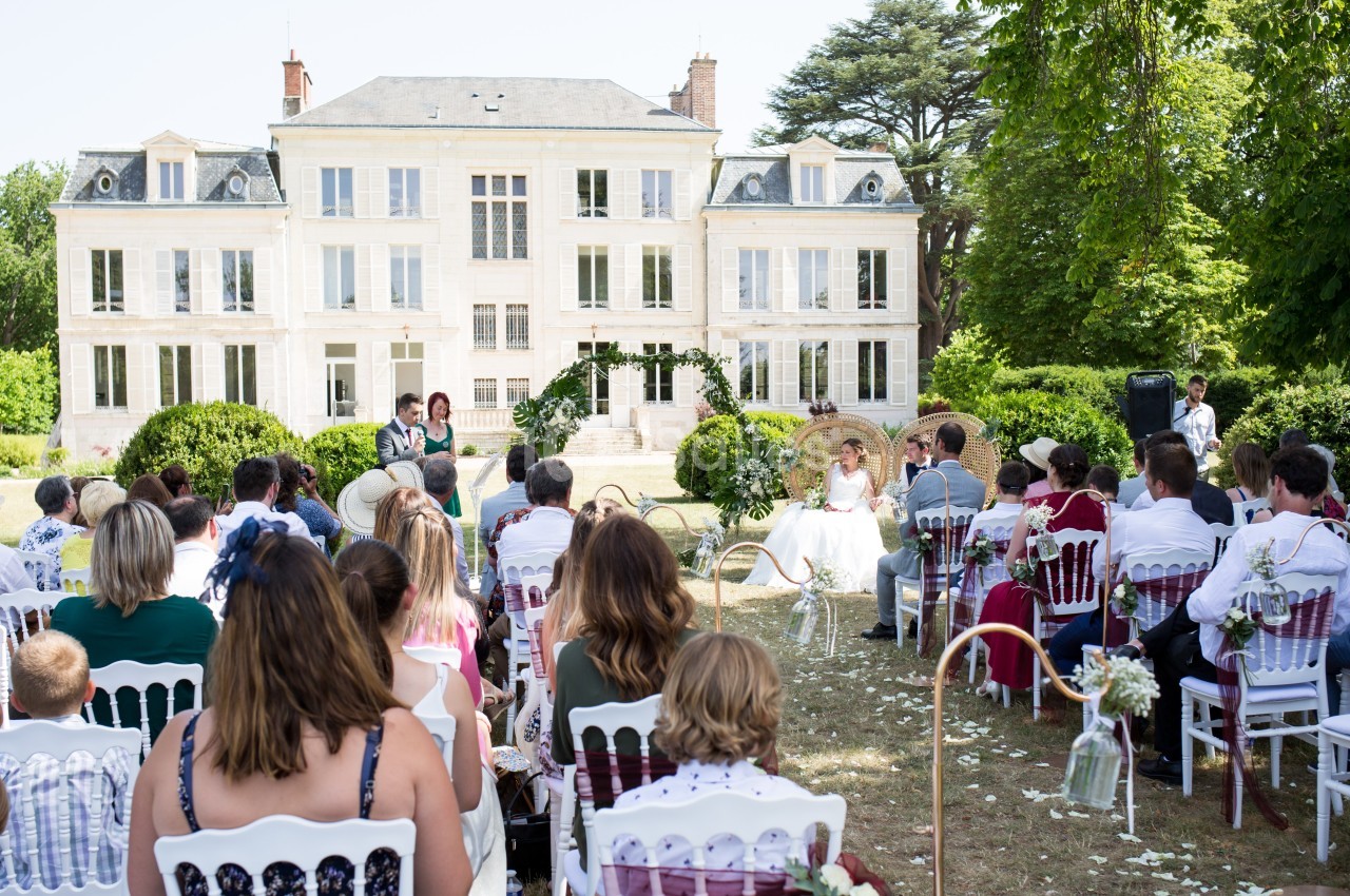 Cérémonie de mariage en extérieur devant un grand bâtiment blanc, avec invités assis et couple au centre.