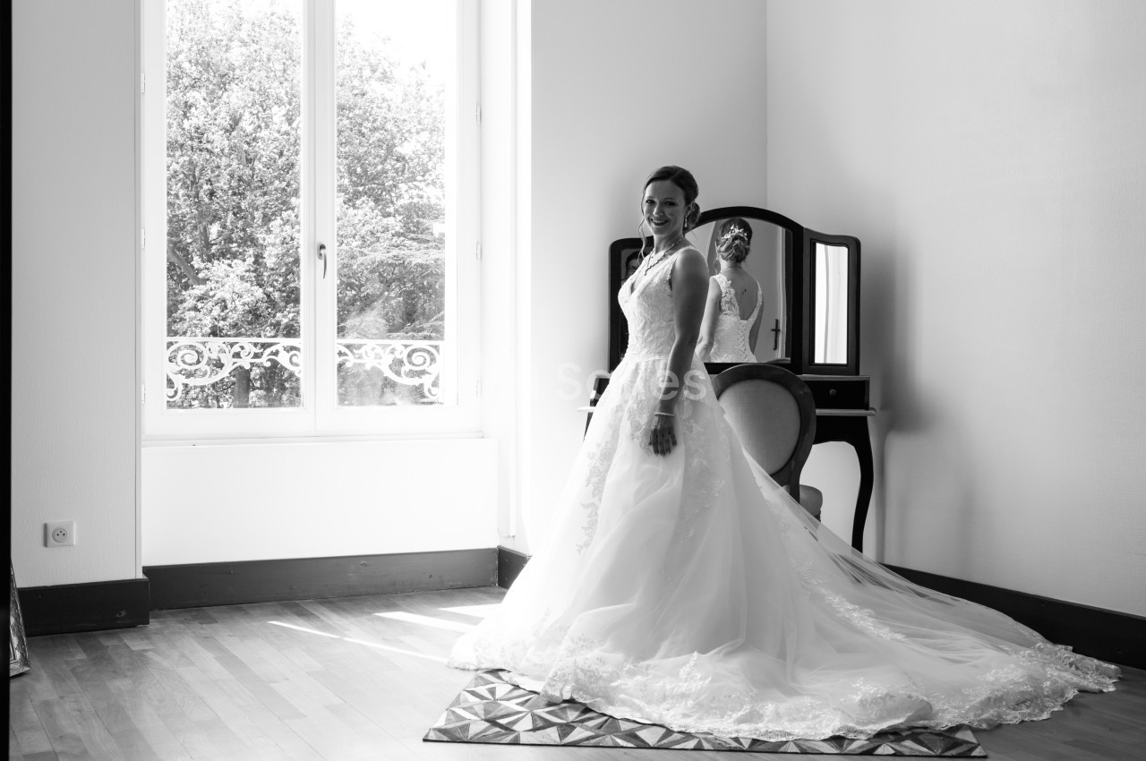 Femme en robe de mariée souriant devant un miroir dans une pièce lumineuse avec une grande fenêtre.