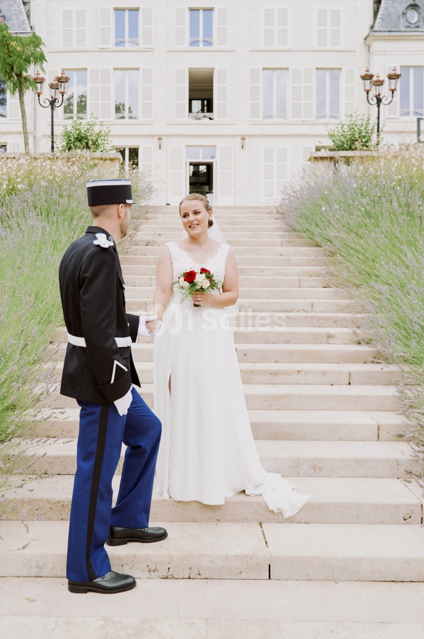 Une mariée en robe blanche tient un bouquet face à un homme en uniforme devant un escalier bordé de lavande.