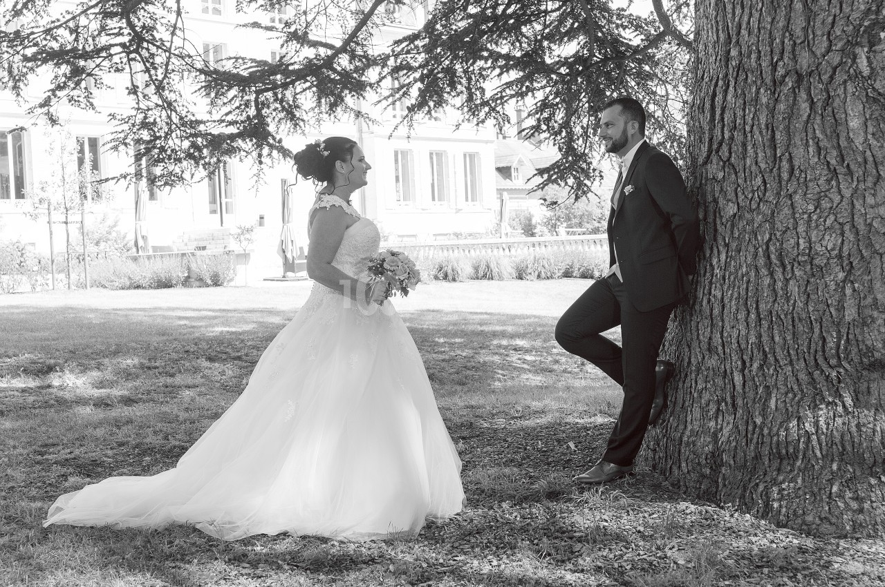 Un couple en tenue de mariage pose sous un arbre dans un parc, la mariée tenant un bouquet.