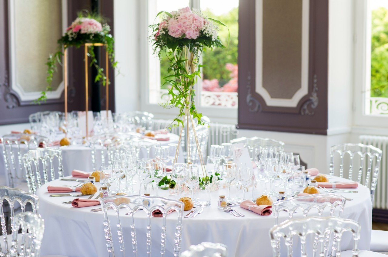 Salle de réception avec tables rondes décorées de nappes blanches, fleurs roses et chaises transparentes.