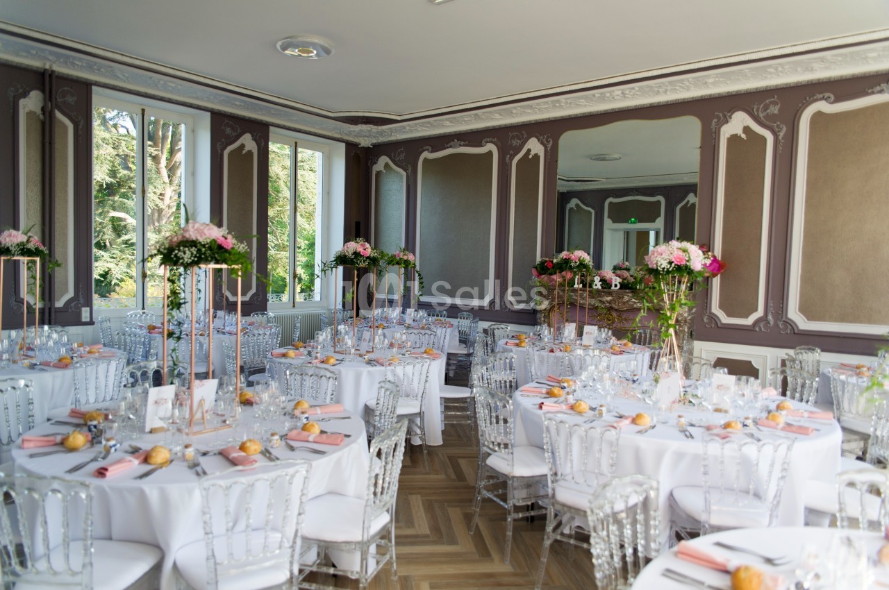 Salle de réception élégante avec tables rondes dressées, décor floral et chaises transparentes.