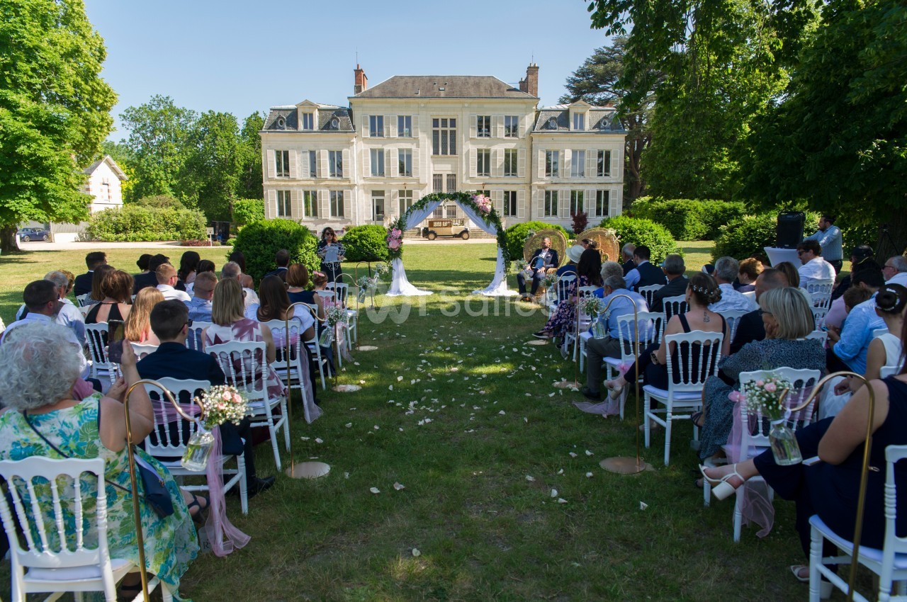 Cérémonie de mariage en plein air devant un manoir, avec des invités assis et une arche décorée de fleurs.