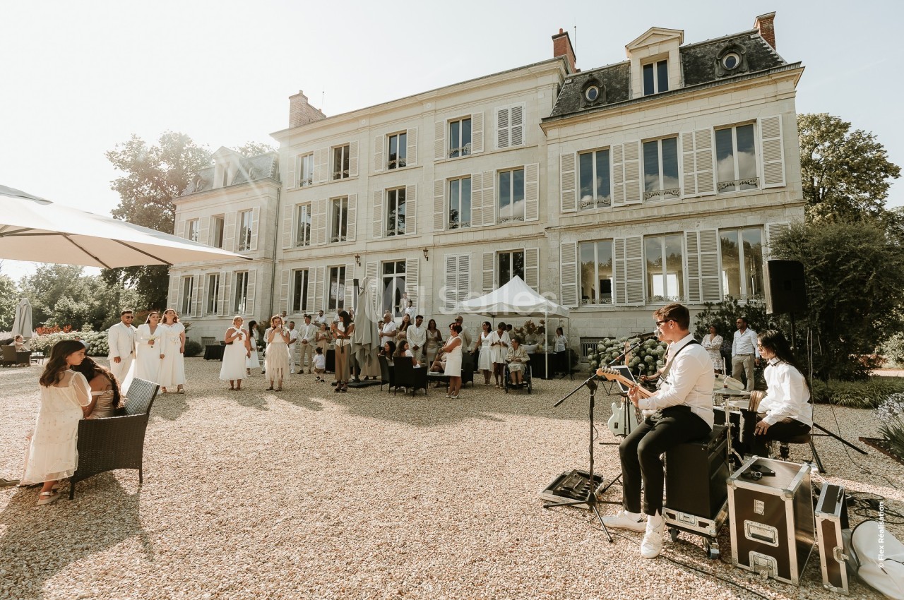 Un groupe de musiciens joue en plein air devant un grand bâtiment de style classique, entouré d'invités en tenue claire.