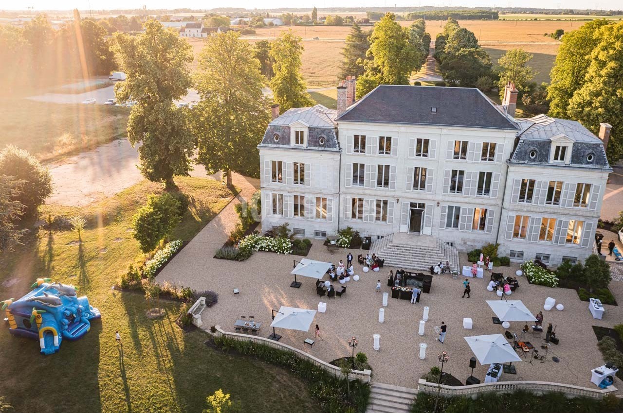 Vue aérienne d'un manoir entouré de jardins, avec une réception en plein air et une structure gonflable pour enfants.