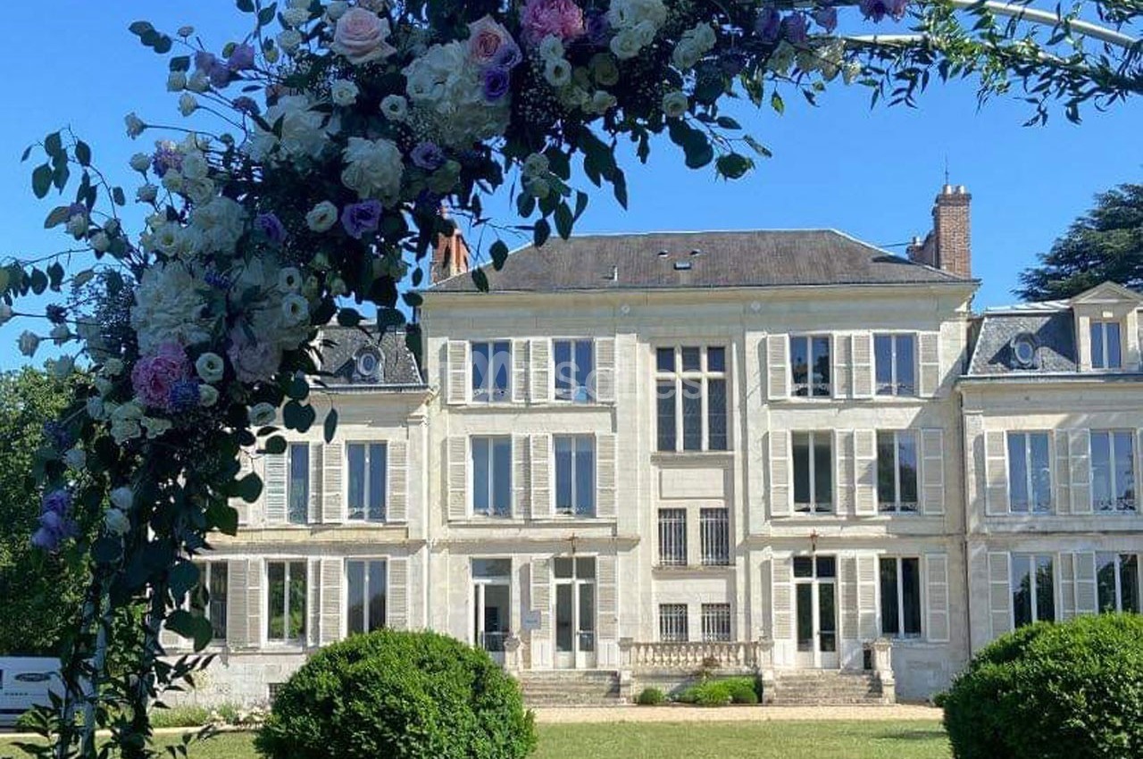 Arche fleurie devant une grande maison de style classique avec façade blanche et jardin verdoyant.