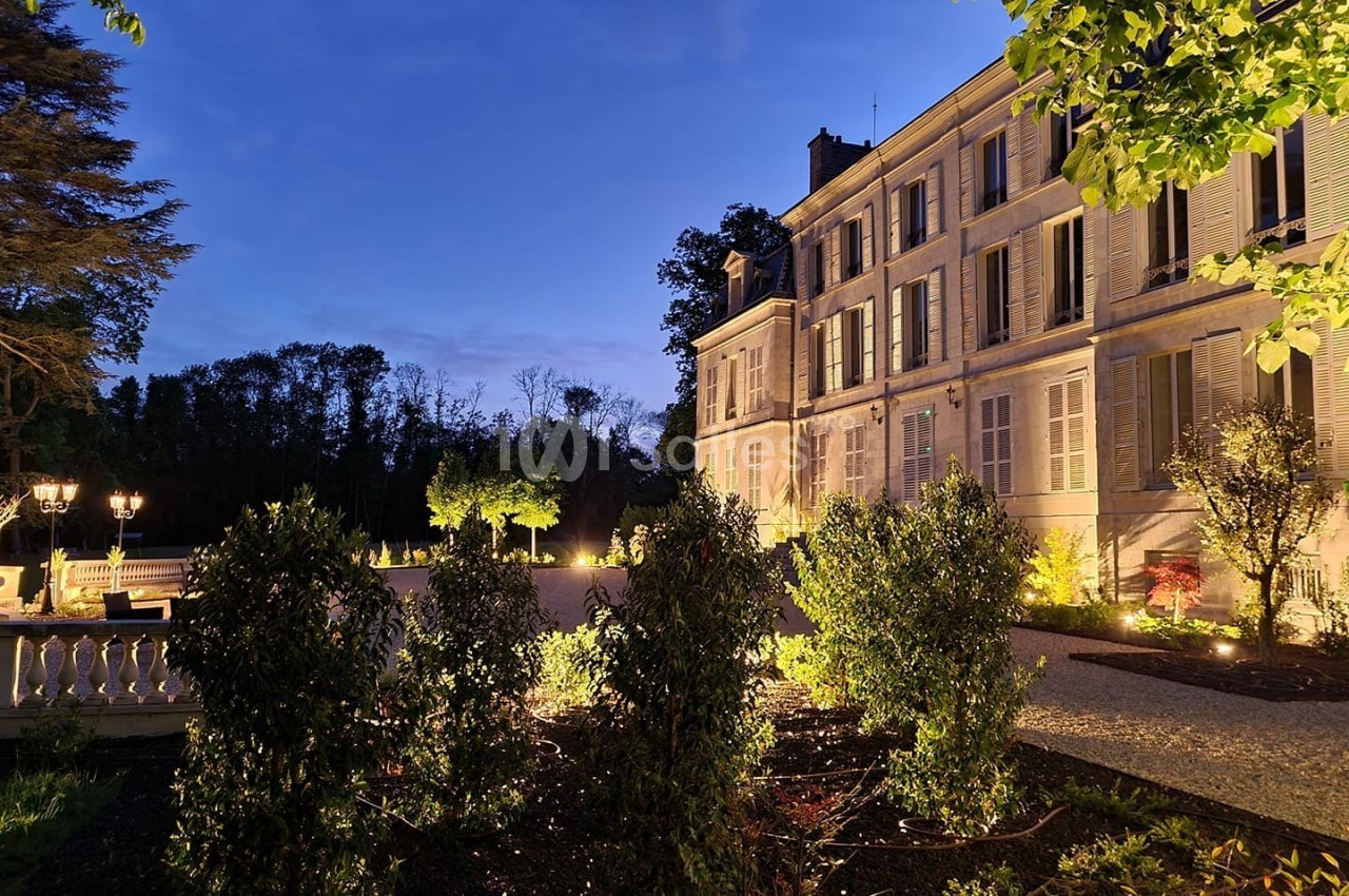Façade d'un bâtiment élégant éclairé au crépuscule, entouré de végétation et d'une allée gravillonnée.