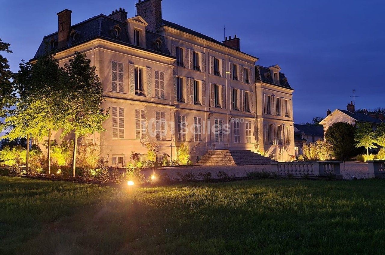 Façade d'un grand bâtiment ancien éclairé la nuit, entouré d'arbres et d'une pelouse.