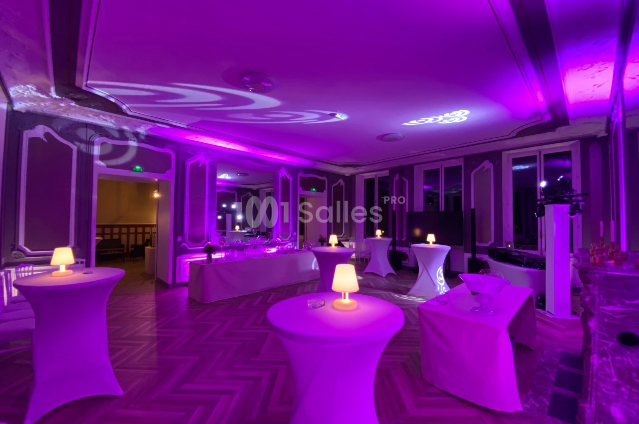 Salle élégante avec éclairage violet, tables hautes nappées de blanc et petites lampes diffusant une lumière douce.