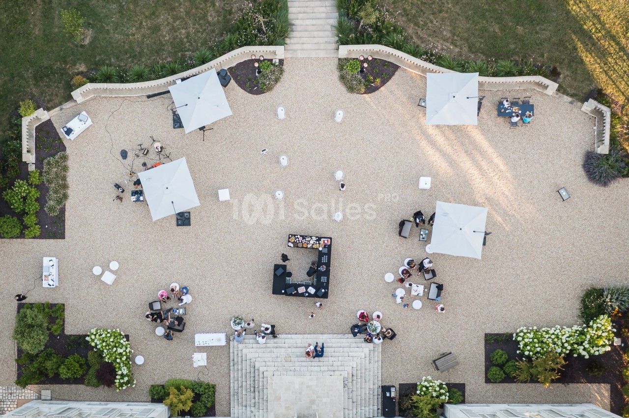 Vue aérienne d'une terrasse avec des tables, des parasols blancs et un bar central entouré de personnes.