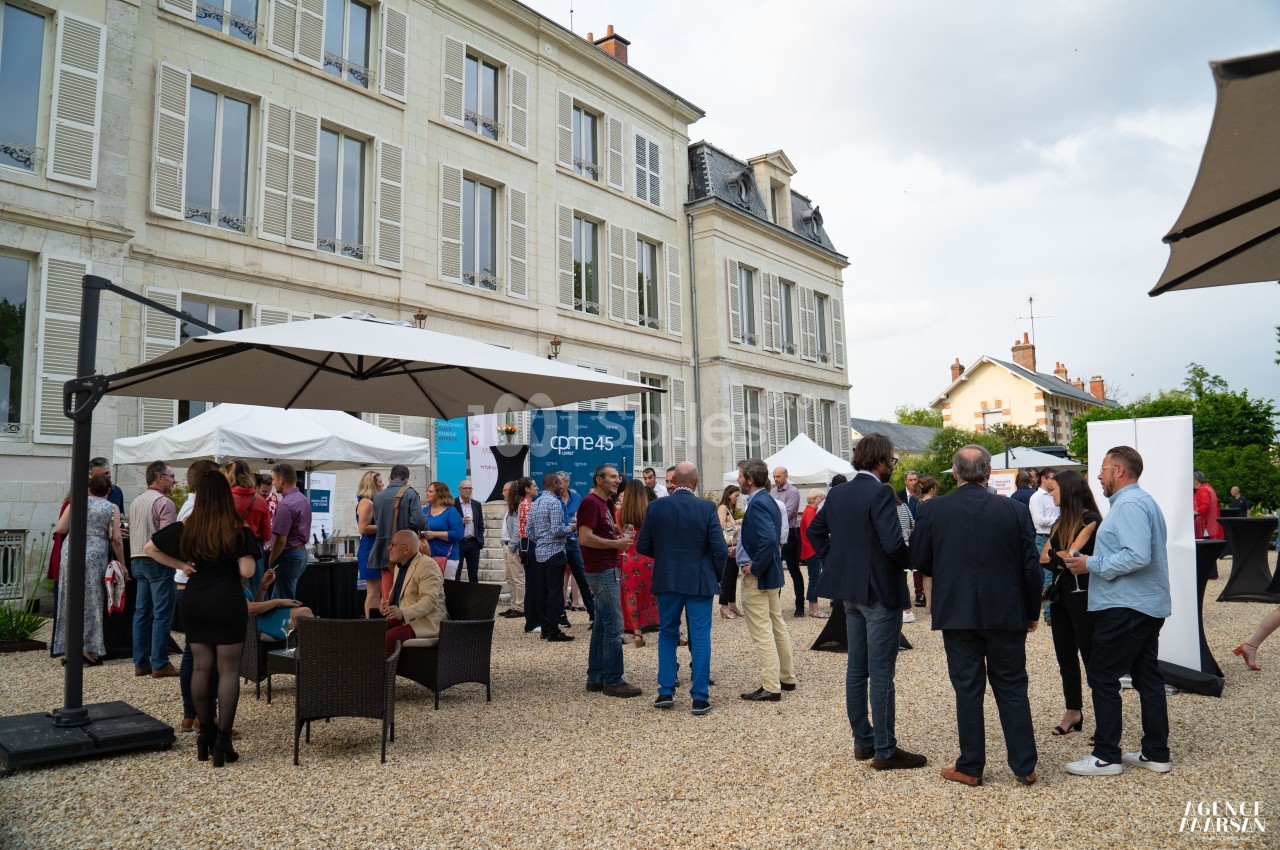 Groupe de personnes discutant dans une cour extérieure devant un bâtiment ancien, sous des parasols et un ciel nuageux.