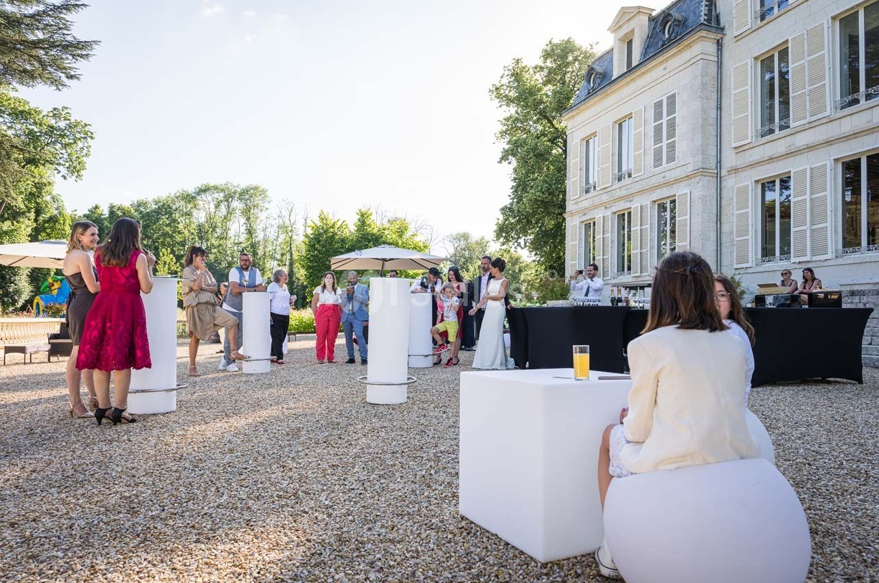 Personnes rassemblées dans une cour gravillonnée devant un bâtiment élégant, discutant autour de tables hautes.