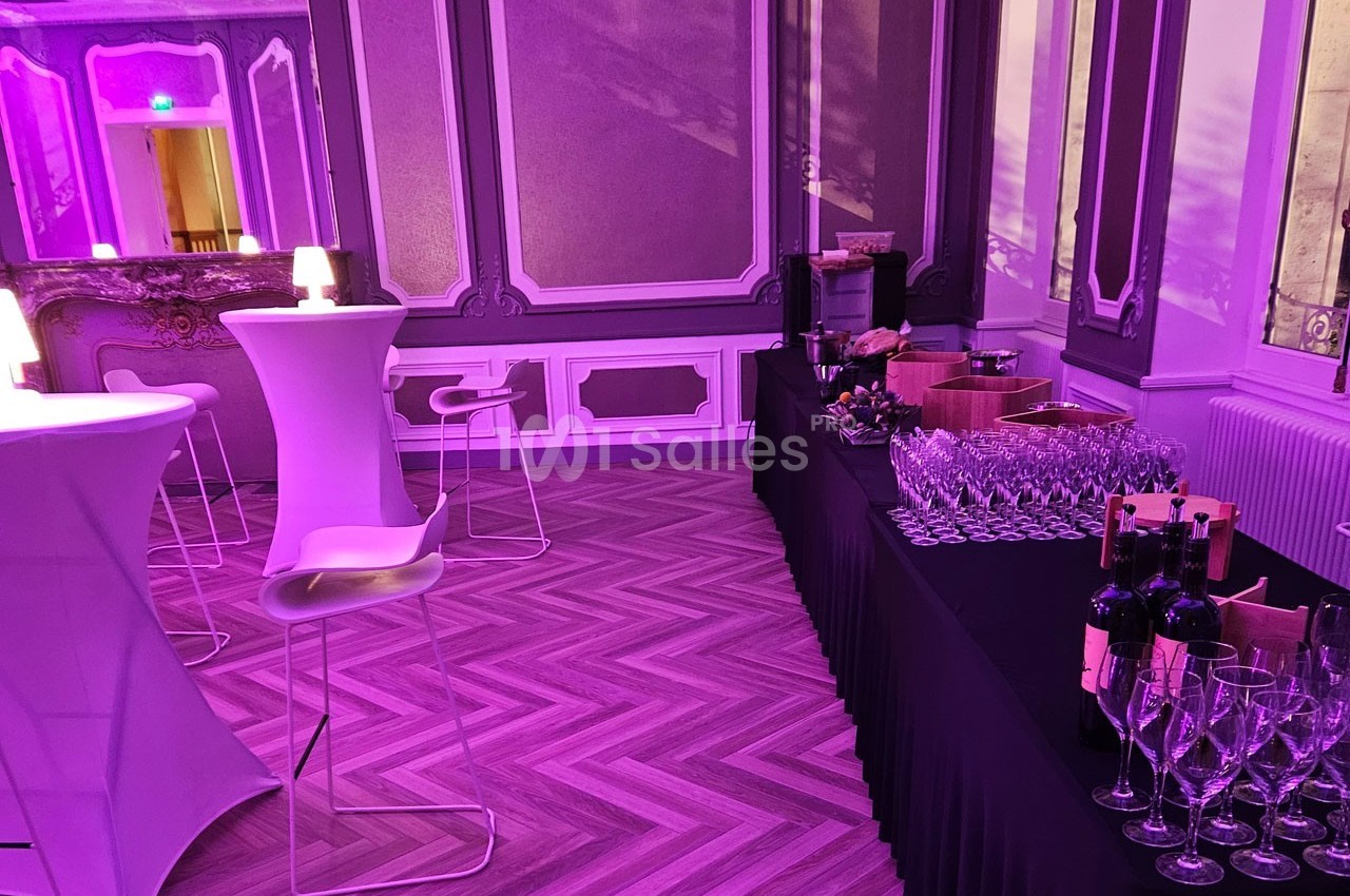 Salle élégante avec tables hautes, chaises modernes et buffet de verres et bouteilles de vin, éclairée par une lumière…