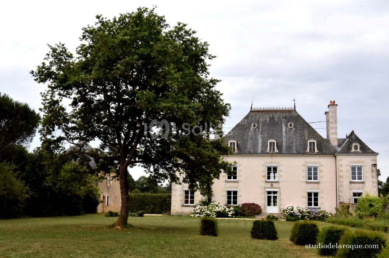 Manoir en pierre blanche avec toit en ardoise, entouré d'un jardin arboré sous un ciel nuageux.