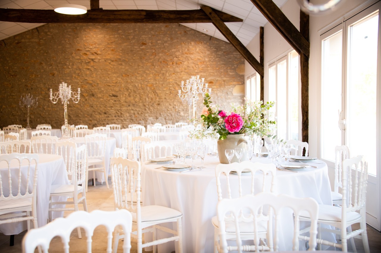 Salle de réception lumineuse avec tables rondes dressées, chaises blanches et décoration florale élégante.