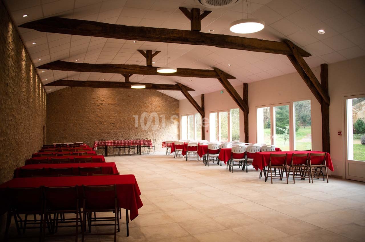 Salle spacieuse avec murs en pierre, poutres apparentes, tables rouges et grandes baies vitrées donnant sur un jardin.