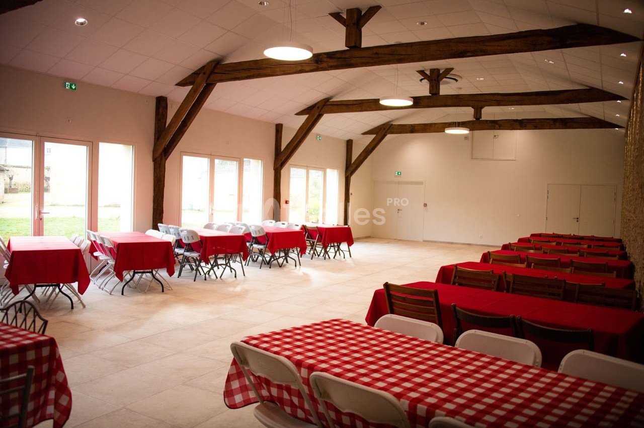 Salle lumineuse avec poutres apparentes, tables rouges et blanches alignées, et grandes baies vitrées donnant sur l…