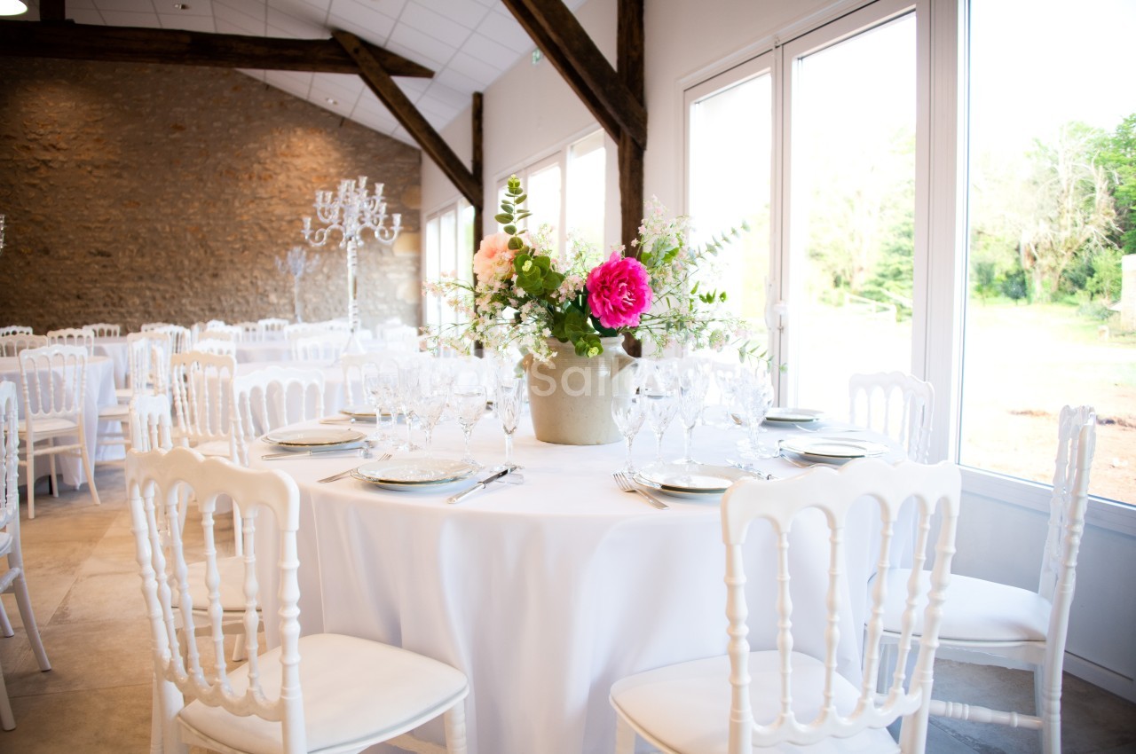 Salle de réception lumineuse avec tables rondes dressées, décorées de fleurs dans un vase central.