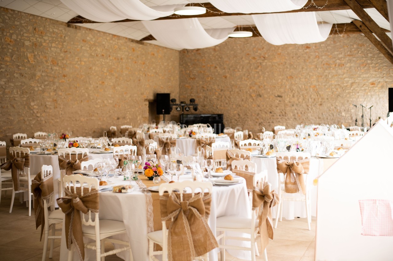 Salle de réception décorée avec des tables rondes, nappes blanches, chaises ornées de nœuds en toile de jute et fleurs…