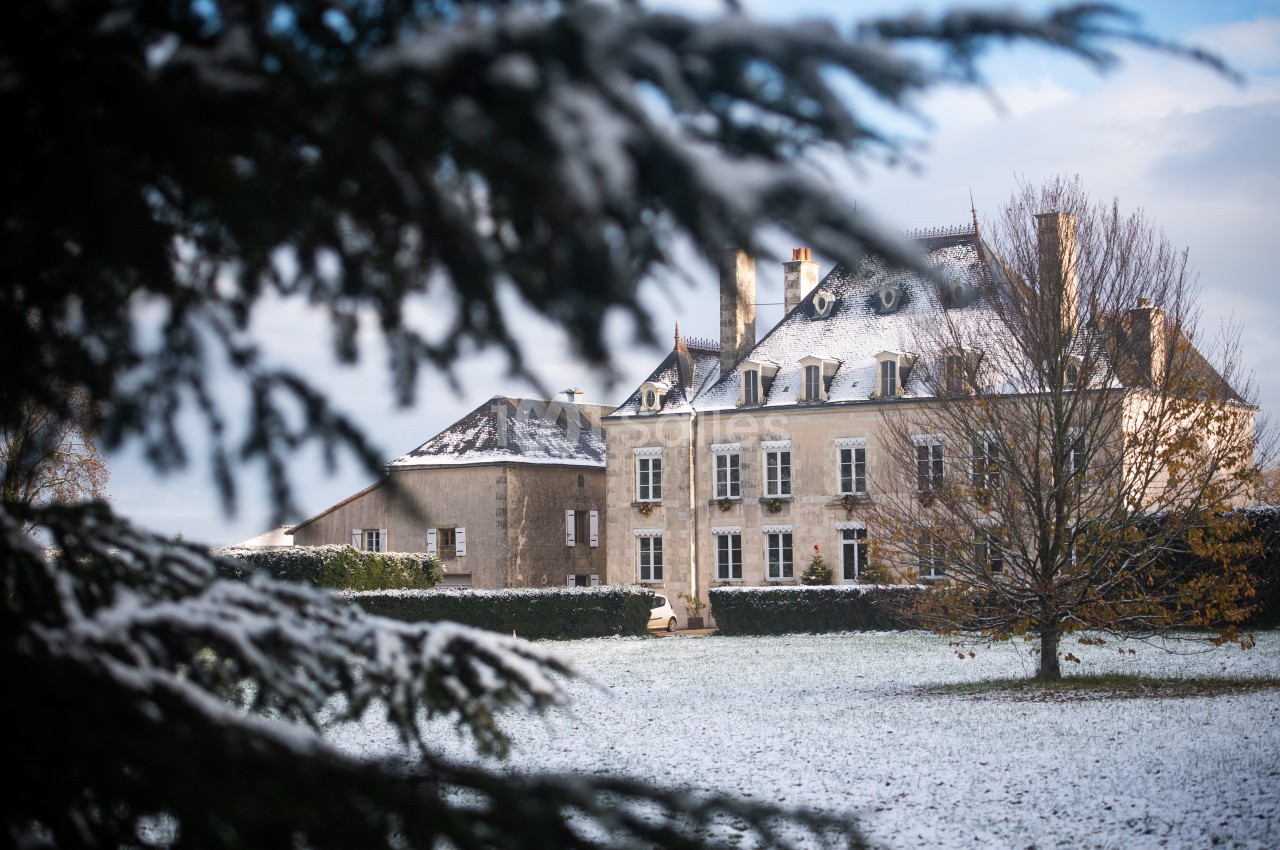 Manoir en pierre entouré d'un jardin enneigé, partiellement encadré par des branches d'arbres au premier plan.