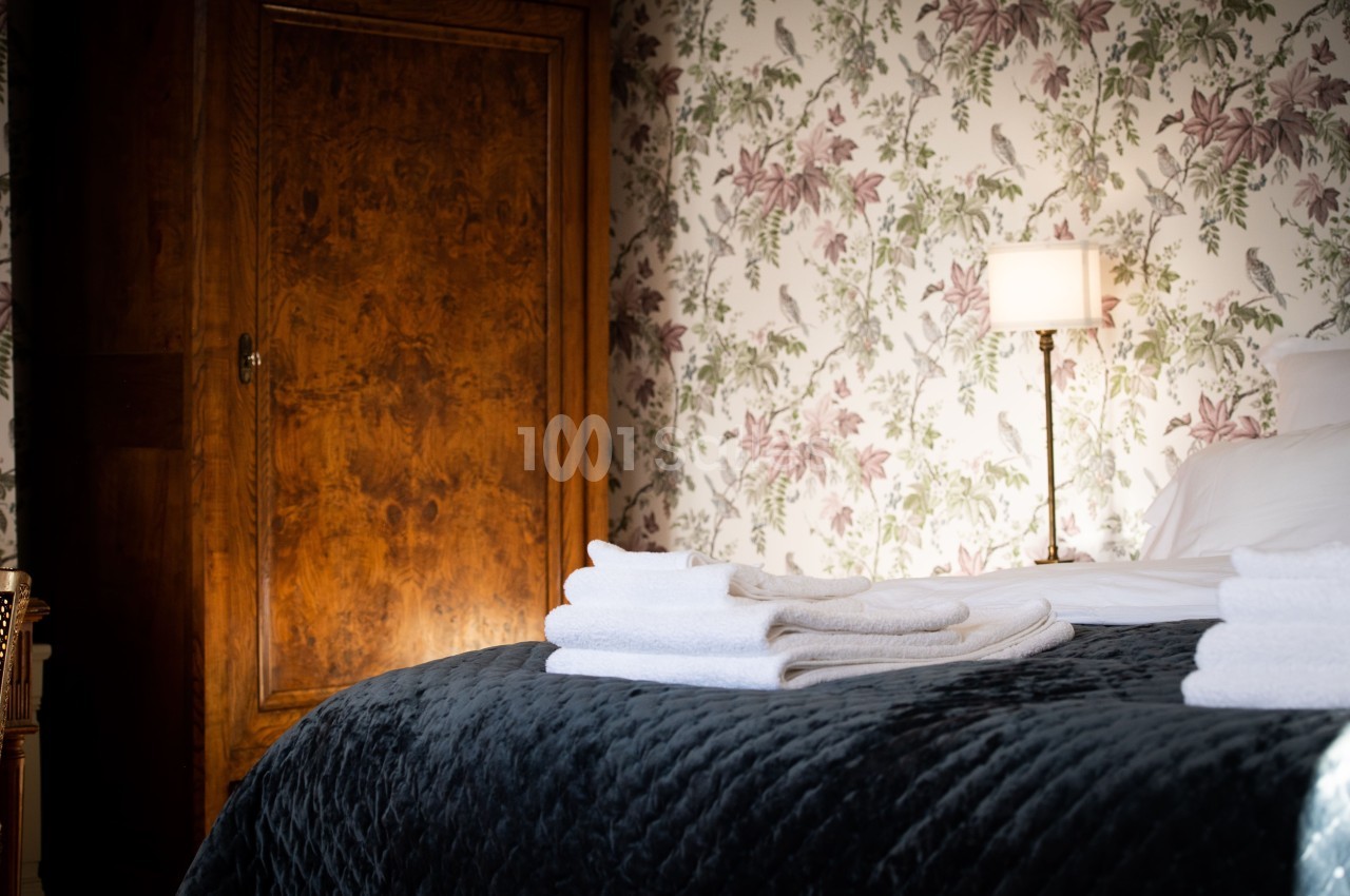 Lit avec couvre-lit sombre, serviettes blanches pliées, armoire en bois et mur tapissé de motifs floraux en arrière-plan.