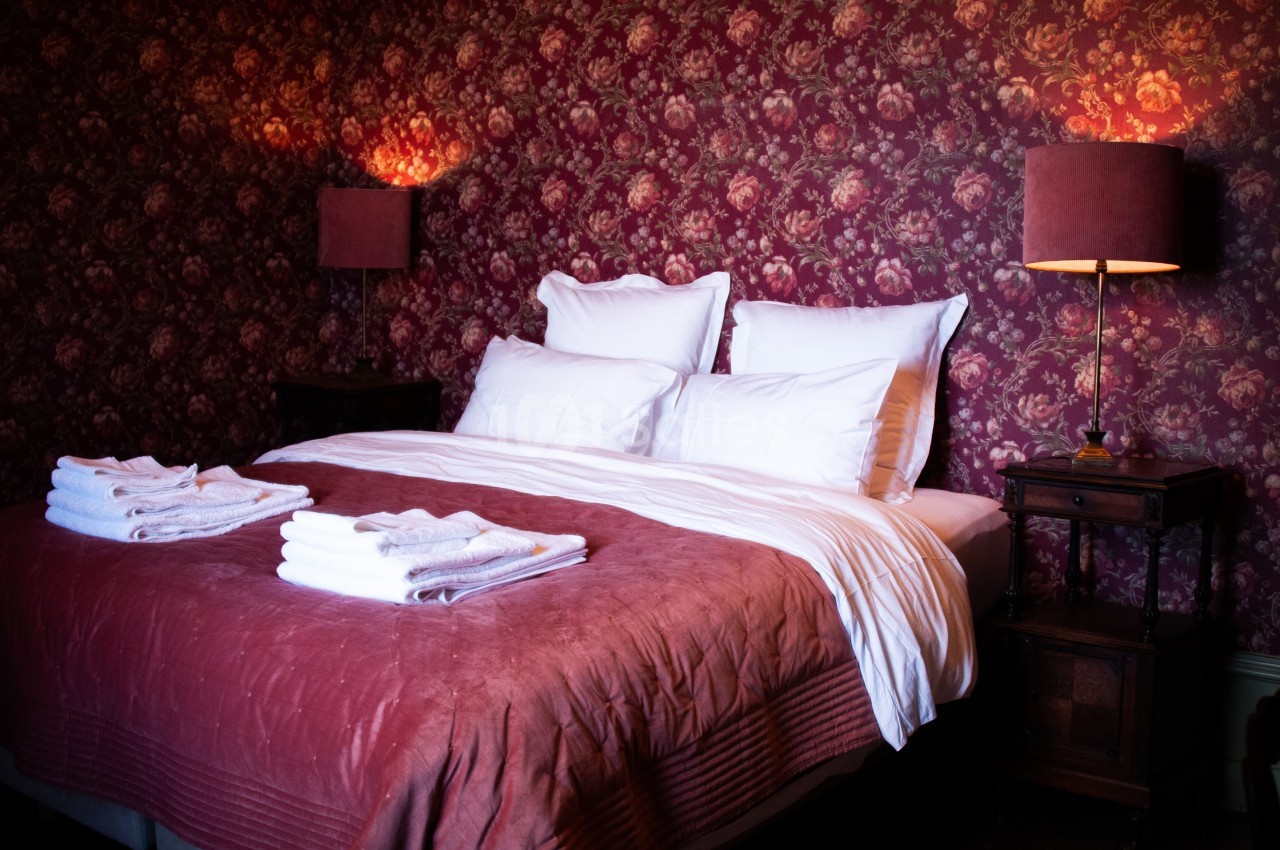 Lit double avec draps blancs, couverture rouge et serviettes pliées, entouré de lampes et d'un papier peint floral.